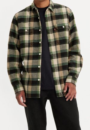 Homme portant une chemise à carreaux verte, beige et noire à manches longues déboutonnée par-dessus un t-shirt noir et un pantalon noir.