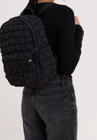 Mochila negra acolchada con un diseño texturizado y abultado, que cuenta con dos bolsillos con cremallera y correas ajustables, usada sobre una camiseta negra y vaqueros.