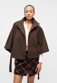 Γυναίκα που φοράει ένα καφέ κάπα μπουφάν πάνω από ένα λευκό cropped πουλόβερ και μια σκούρα κόκκινη και μαύρη καρό φούστα με κουμπιά, στέκεται μπροστά σε απλό φόντο.