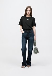 Juoda medvilninė marškinėliai su logotipu, tamsiai mėlyni platėjančių kelnių džinsai ir žalia tekstūruota rankinė. Modelis dėvi šlepetes ir stovi neutraliame fone.