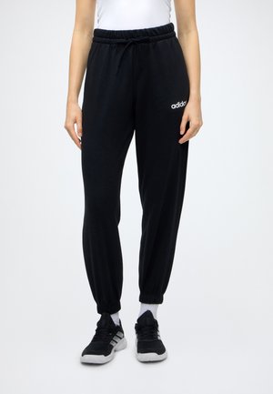 Persona que lleva pantalones jogger Adidas negros con puños y cinturilla elásticos, calcetines blancos, zapatillas negras y blancas, de pie sobre un fondo blanco.