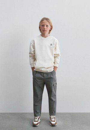 Niño con cabello rubio lleva sudadera color crema, pantalones cargo grises y zapatillas blancas y marrones, de pie frente a un fondo claro y liso.