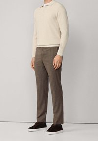 Homme portant un pull polo manches longues crème, un pantalon à fines rayures marron et des baskets marron foncé à semelles blanches, debout sur un fond neutre.