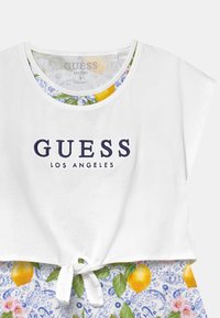 Valkoinen crop-top, jossa on "GUESS LOS ANGELES" -logo laivastonsinisellä. Ominaisuuksiin kuuluu solmittu etuosa ja kukkakuosiset helmavarat, joissa on sitruunoita ja sinisiä yksityiskohtia.