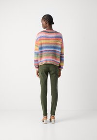 Vrouw met gevlochten haar, gekleed in een gestreepte trui met meerdere kleuren, olijfgroene broek en witte hakken, staand tegen een plain witte achtergrond.