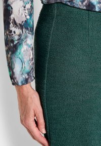 Groene gestructureerde rok van een zachte, verheven stof. Een hand houdt de zijnaad van de rok vast. De top heeft een kleurrijk bloemenpatroon.
