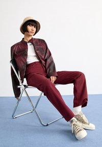 Bordowa skórzana kurtka, biała bluzka, bordowe spodnie o wysokim stanie, białe skarpetki typu crew i sneakersy w zebrę. Modelka siedzi na metalowym krześle.