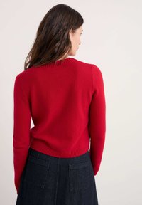 Sarkans adīts džemperis ar ribotu kakla izgriezumu un apakšmalu, pieguļošs siluets, skatīts no aizmugures uz neitrāla fona, kombinēts ar tumšu džinsa svārku.