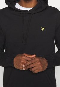 Felpa nera in misto cotone, con cappuccio regolabile tramite cordino, polsini a coste e un piccolo logo giallo a forma di aquila sul petto.