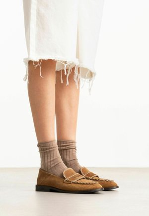 Onderbenen met beige suède loafers met witte steken en bruine geribbelde sokken, gecombineerd met witte cropped broeken met een rafelige zoom.