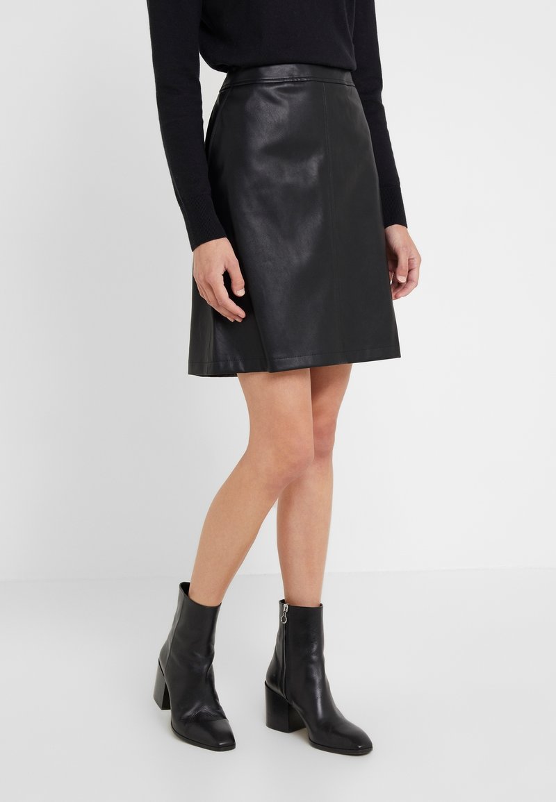 Jupe en cuir noire évasée portée avec un haut noir à manches longues et des bottines noires à talons sur une personne debout.