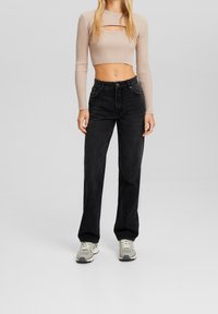 Top corto de canalé en beige claro con mangas largas y corte en el pecho, combinado con pantalones vaqueros de pierna ancha negros de tiro alto y zapatillas grises.