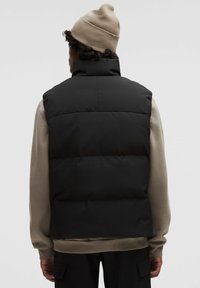 Schwarze gepolsterte Weste mit hohem Kragen, von hinten dargestellt. Trägt man über einem hellgrauen Sweatshirt, ergänzt durch eine beigen Strickmütze.