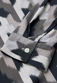Camicia button-up con un motivo astratto in nero, grigio e bianco. Presenta un tessuto morbido, un colletto a punta e un solo bottone bianco sul polsino.