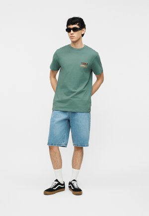 Fiatal férfi zöld mintás pólóban, világos kék farmer rövidnadrágban, fekete tornacipőben fehér zoknival és fekete napszemüvegben, kezét a háta mögött tartva áll.