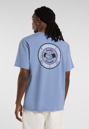 Lyseblå bomulds t-shirt med et cirkulært grafisk motiv på ryggen, der viser et håndtryk, sort og rød tekst samt korte ærmer.