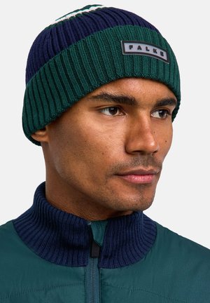 Berretto lavorato a maglia con un design a strisce verde e blu navy, caratterizzato da una texture a coste e una toppa con logo. Indossato con una giacca abbinata a colletto alto.
