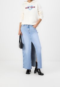 Persona vestida con una sudadera blanca de "Nueva York", una falda larga de mezclilla azul claro con una abertura frontal, botas de tacón negras y sosteniendo un pequeño bolso negro.