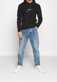 Felpa nera con un logo bianco, abbinata a jeans in denim azzurro chiaro e sneakers bianche. L'outfit combina elementi casual e confortevoli.