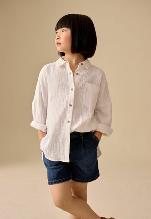 Child with short black hair standing with hands in pockets, wearing a white button-up shirt and dark denim shorts against beige background.
