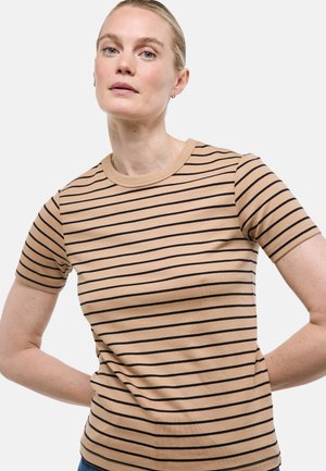 Femme aux cheveux blonds attachés, portant une chemise beige à manches courtes avec des rayures horizontales noires, debout devant un fond uni.