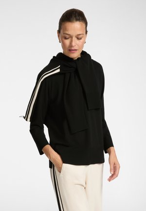 Schwarzes Strickoberteil mit Kapuze, mit weißen gestreiften Akzenten an den Schultern und gerippten Bündchen. Kombiniert mit cremefarbenen Hosen.