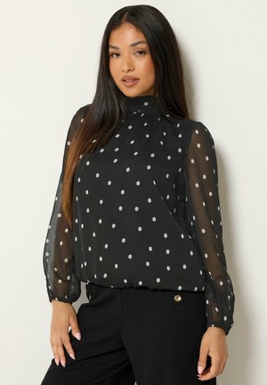 Jeune femme aux longs cheveux foncés portant un chemisier noir transparent à pois et un pantalon noir sur un fond clair uni.