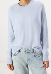 Person wearing a light blue crewneck sweater and light wash denim jeans, hands relaxed at sides against a plain background.