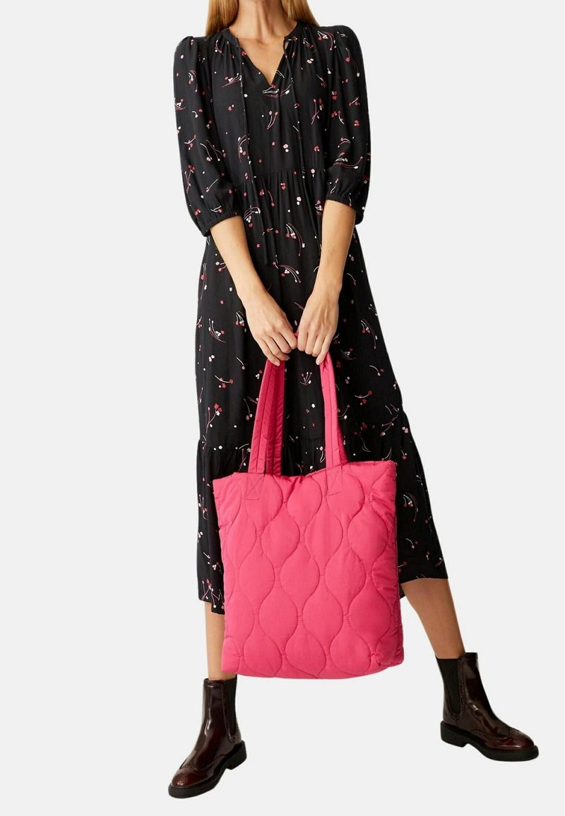 Femme portant une robe midi noire à fleurs et des bottines foncées, tenant un grand sac cabas matelassé rose avec deux poignées.
