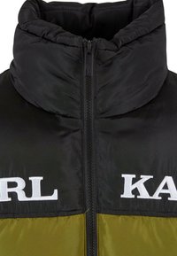 Puffjakke med en høj, quiltet krave. Har sort øverste halvdel og olivengrøn nederste halvdel. Stor hvid "KARL" logo på fronten. Lynlås lukning.