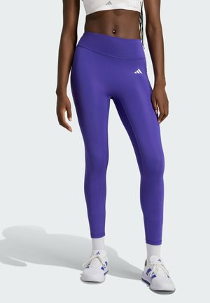 Frau trägt violette Adidas-Leggings, weißen Sport-BH, weiße Adidas-Sneakers und steht vor einem schlichten hellen Hintergrund.