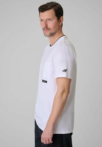 Homme aux cheveux courts et à la barbe portant un t-shirt blanc de la marque 4F avec une poche poitrine, debout de profil devant un fond gris uni.