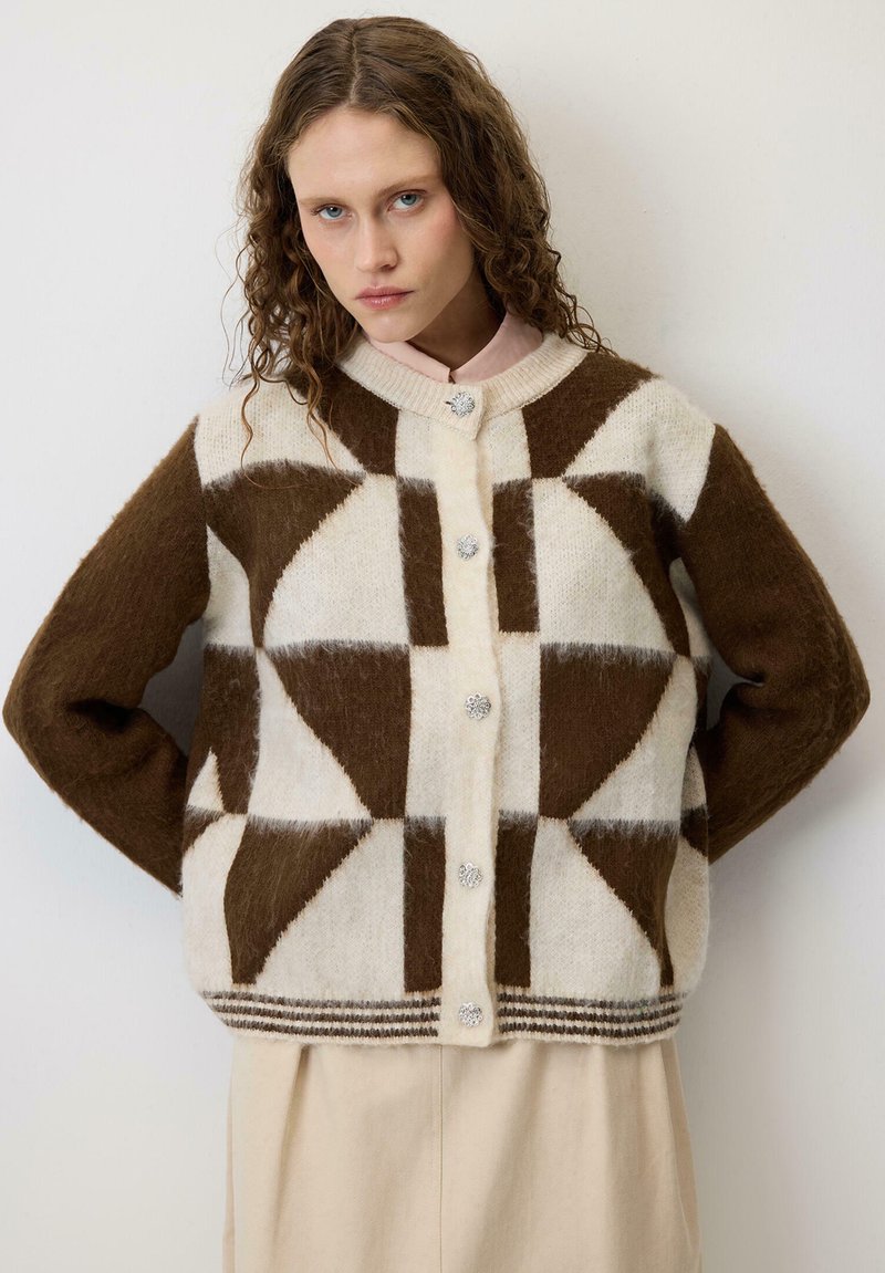 Γυναίκα με σγουρά μαλλιά που φοράει καφέ και κρεμ ζακέτα με γεωμετρικό μοτίβο και μπεζ φούστα, στέκεται μπροστά σε έναν λευκό τοίχο.