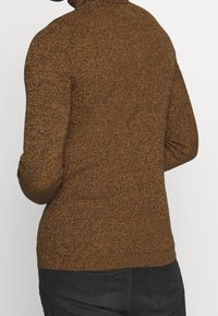 Personne portant un pull en maille texturée brun et un pantalon noir, vue de dos, sur un fond clair uni.