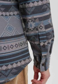 Hosszú ujjú ing, geometrikus mintával szürke, kék és fekete árnyalatokban. Texturált anyag, gombos mandzsetta és ívelt szegély részletek.