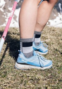 Bottes de randonnée Millet bleu clair avec une tige texturée, des accents noirs et des semelles blanches, associées à des chaussettes rayées grises. Le sol est herbeux.