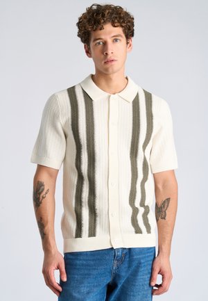 Jonge man met krullend haar die een crème kleurig overhemd met korte mouwen draagt, voorzien van verticale donkere strepen, en een blauwe spijkerbroek, staand tegen een effen achtergrond.