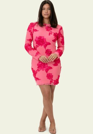 Junge Frau trägt ein pinkes Langarm-Minikleid mit roter Blumenstickerei, steht mit gekreuzten Beinen und verschränkten Händen vorne.