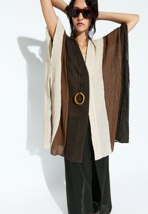 Femme portant un haut oversize bicolore marron, beige et noir avec de larges manches, pantalon noir, lunettes de soleil et collier avec pendentif circulaire.