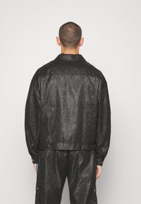 Svart, glänsande jacka gjord av texturerat material med en avslappnad passform, med en krage, långa ärmar och en justerbar nederkant.