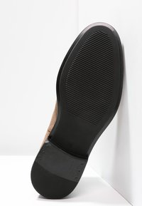 Zapato de ante marrón con suela de goma negra, que presenta un patrón estriado para mayor agarre. El zapato tiene una puntera redondeada y un diseño de perfil bajo.