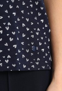 Camiseta de color azul marino con patrones de corazones blancos, mangas cortas, dobladillo redondeado y un pequeño detalle de botón azul cerca de la parte inferior.