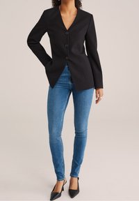 Svart skräddarsydd blazer med djup V-ringning, tre knappar och långa ärmar, kombinerad med blå skinny jeans och svarta spetsiga klackskor.