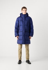 Giacca a vento blu navy con cappuccio, caratterizzata da un design trapuntato, chiusura con zip e tasche laterali. Indossata sopra pantaloni beige e scarpe nere.