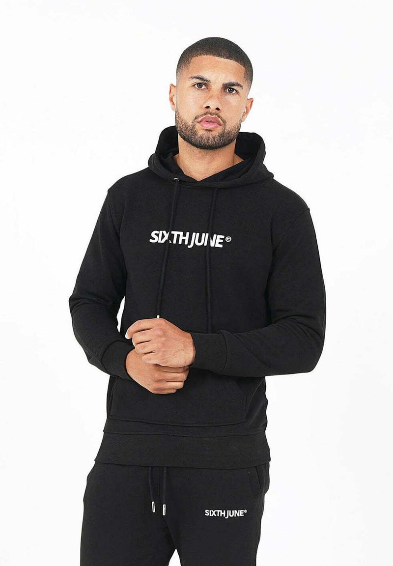 Sudadera de algodón negra con bolsillo canguro, capucha con cordón y logo blanco "SIXTHJUNE®" en la parte frontal. Combinada con pantalones de chándal negros.
