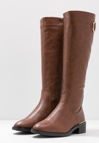 Bottes montantes marron en cuir synthétique, avec un bout arrondi, un talon bloc bas et un accent décoratif en métal sur la tige.