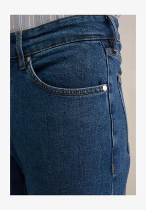 Mørkeblå denimjeans med en glat overflade, der har en forlomma, metalknapdetaljer og kontrastsyninger langs sømmene.