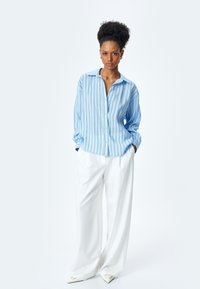 Camisa de botones a rayas azul claro con mangas largas, combinada con pantalones anchos blancos y zapatos puntiagudos blancos. Textura de tela suave.