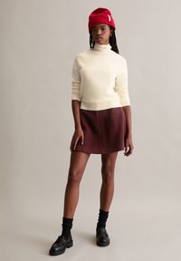 Cremfarbener Rollkragenpullover, burgunderfarbener Filzrock, schwarze Plateau-Loafers und roter Strickmütze. Strukturierten Stoffe mit kontrastierenden Farben.