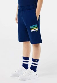 Bambino che indossa pantaloni corti Lacoste blu navy con logo multicolore, calzini a righe blu e bianche e sneakers bianche con velcro.